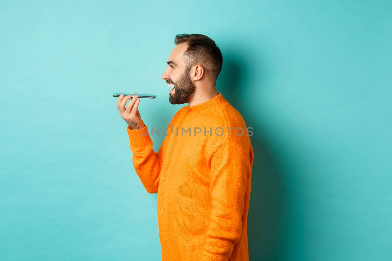 Profile of handsome bearded man recording voice message, holding mobile phone and talking on speakerphone, smiling happy, standing against turquoise background.