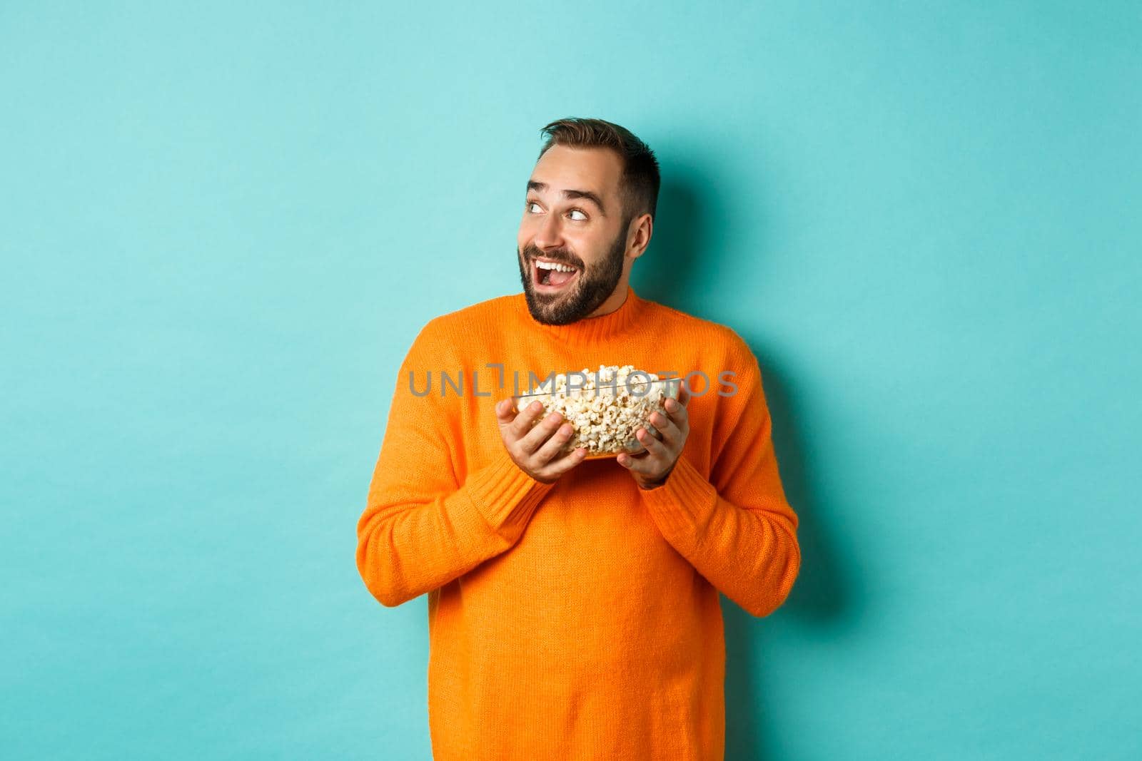 Excited and happy man watching tv and holding bowl of popcorn, looking left and smiling pleased, standing over blue background.