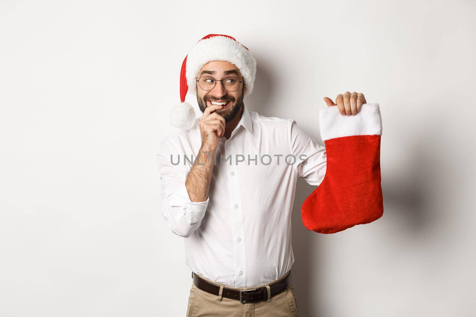 Merry christmas, holidays concept. Adult man looking happy and curious at xmas sock, receive gifts, wearing santa hat, white background.