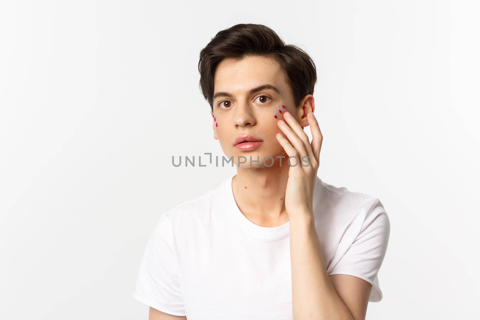 People, lgbtq community and lifestyle concept. Beautiful young gay man applying glitter under eyes for pride party, standing over white background.