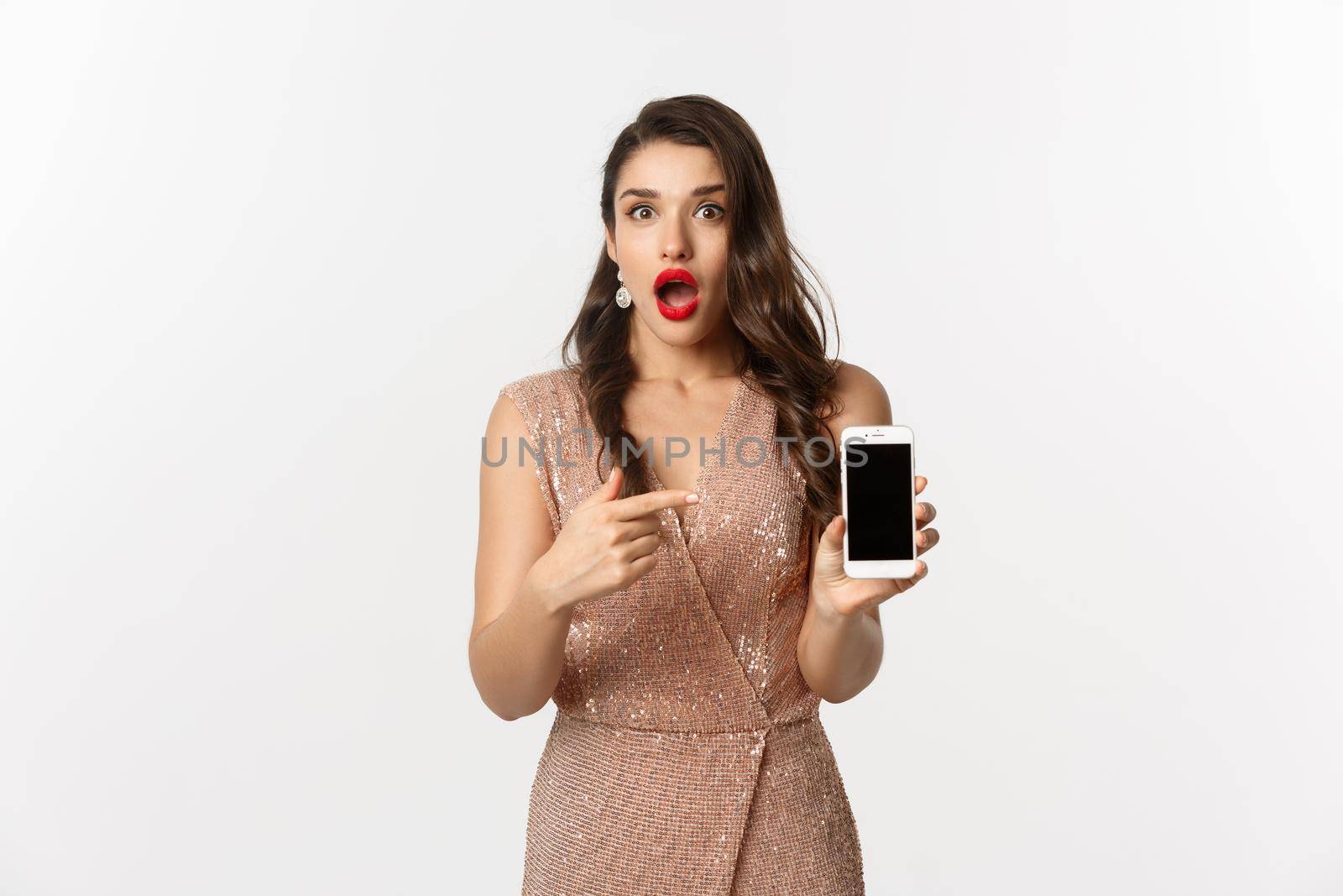 Online shopping. Amazed woman in elegant dress, red lips, pointing finger at mobile screen, showing app, standing over white background.