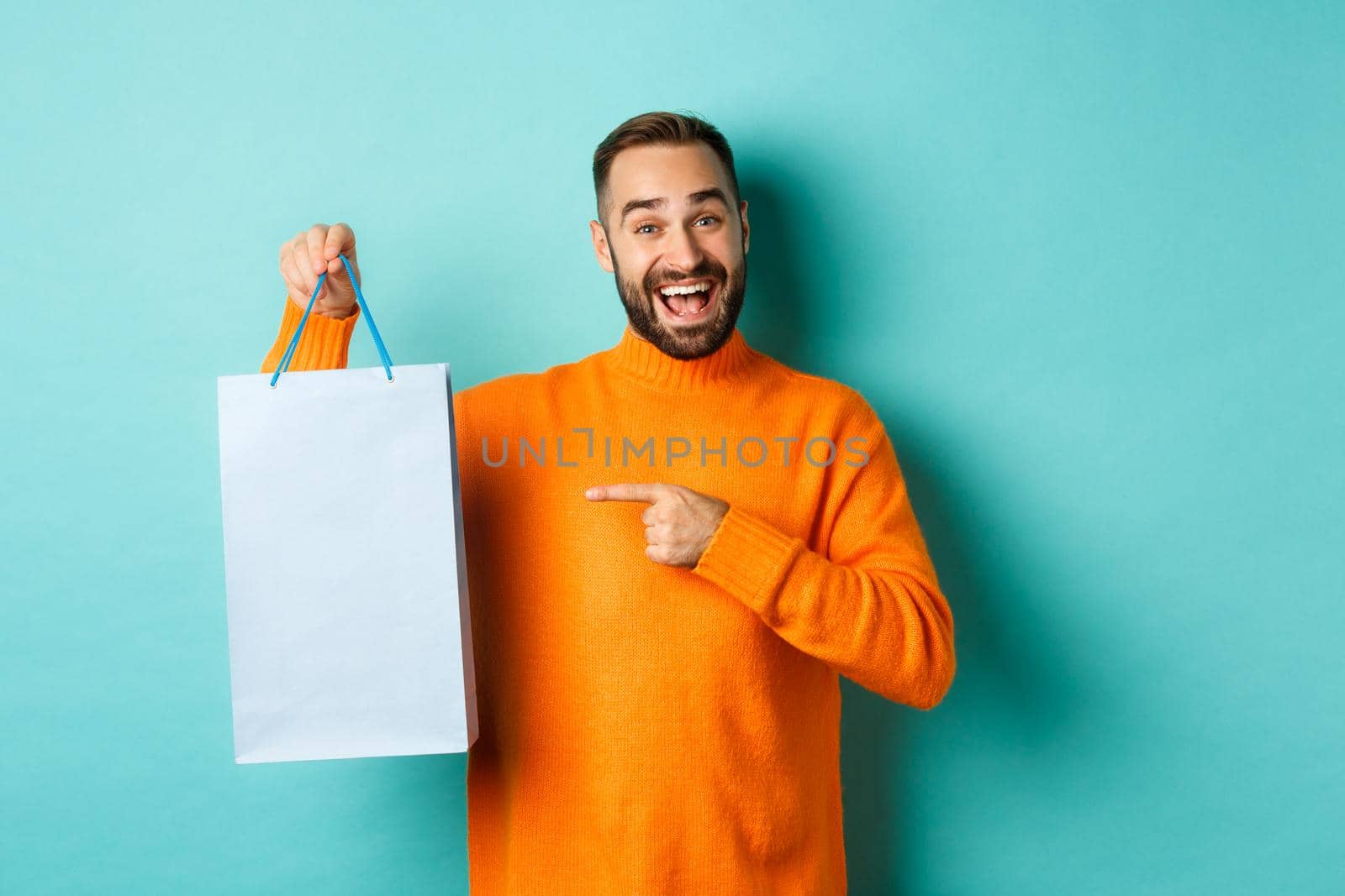 Handsome smiling man pointing finger at shopping bag, buying in stores, standing satisfied over blue background.