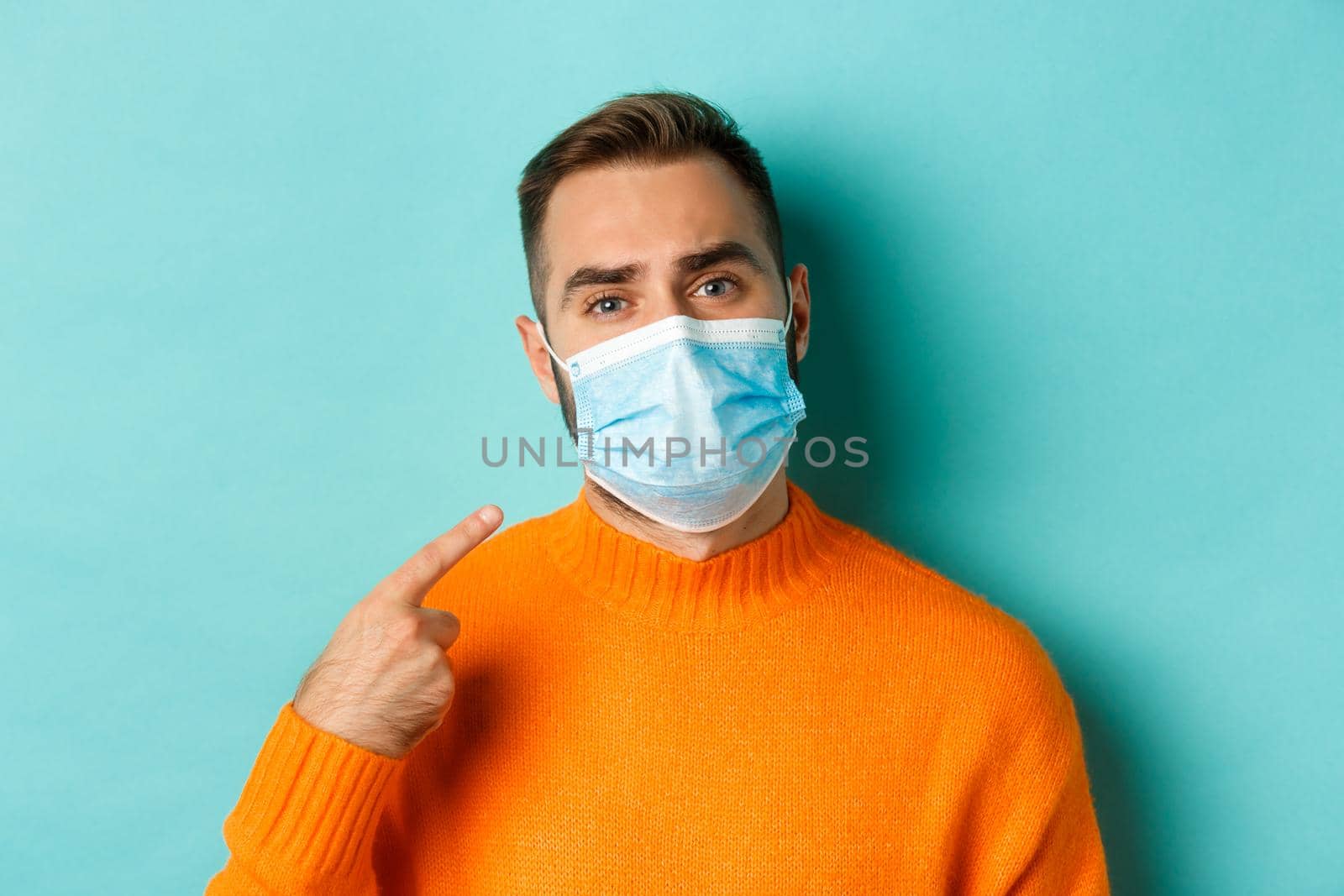 Covid-19, social distancing and quarantine concept. Upset man in complaining on face mask, pointing at it and looking bothered, standing over light blue background.