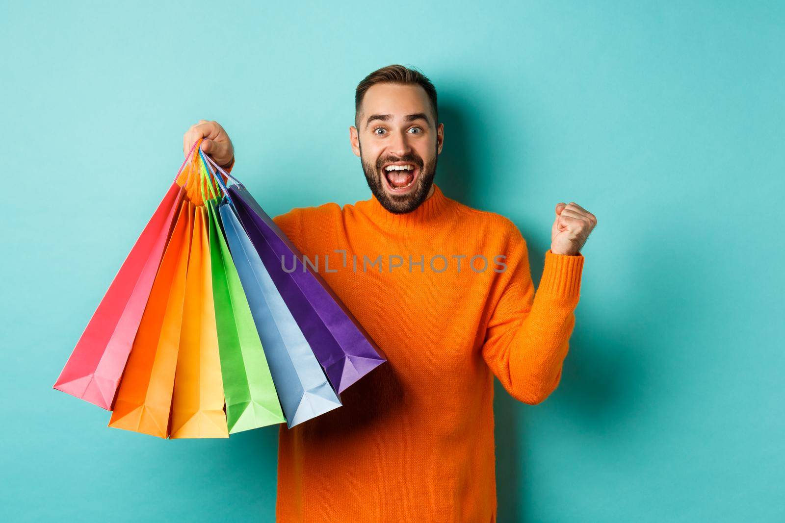 Happy handsome man holding shopping bags and smiling, feeling excitement from discounts, making fist pump, standing over turquoise background.