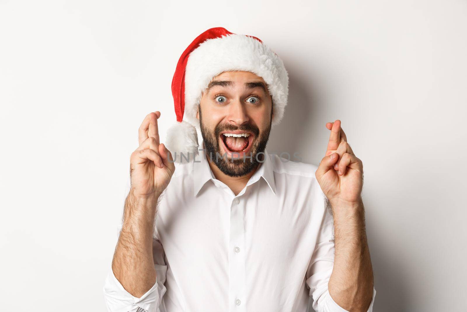 Party, winter holidays and celebration concept. Happy man in santa hat making christmas wish, cross fingers for good luck and looking excited, white background by Benzoix