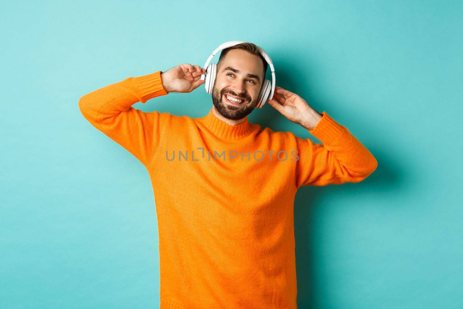 Handsome bearded man listening to music in headphones, smiling pleased, standing over turquoise background.