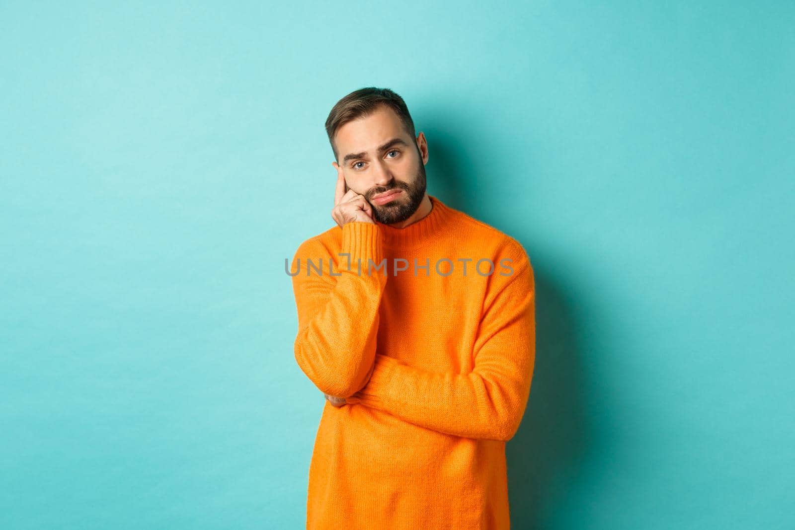 Sad and gloomy man with beard, looking indesicive and sad, thinking about something and sulking, standing over turquoise background.