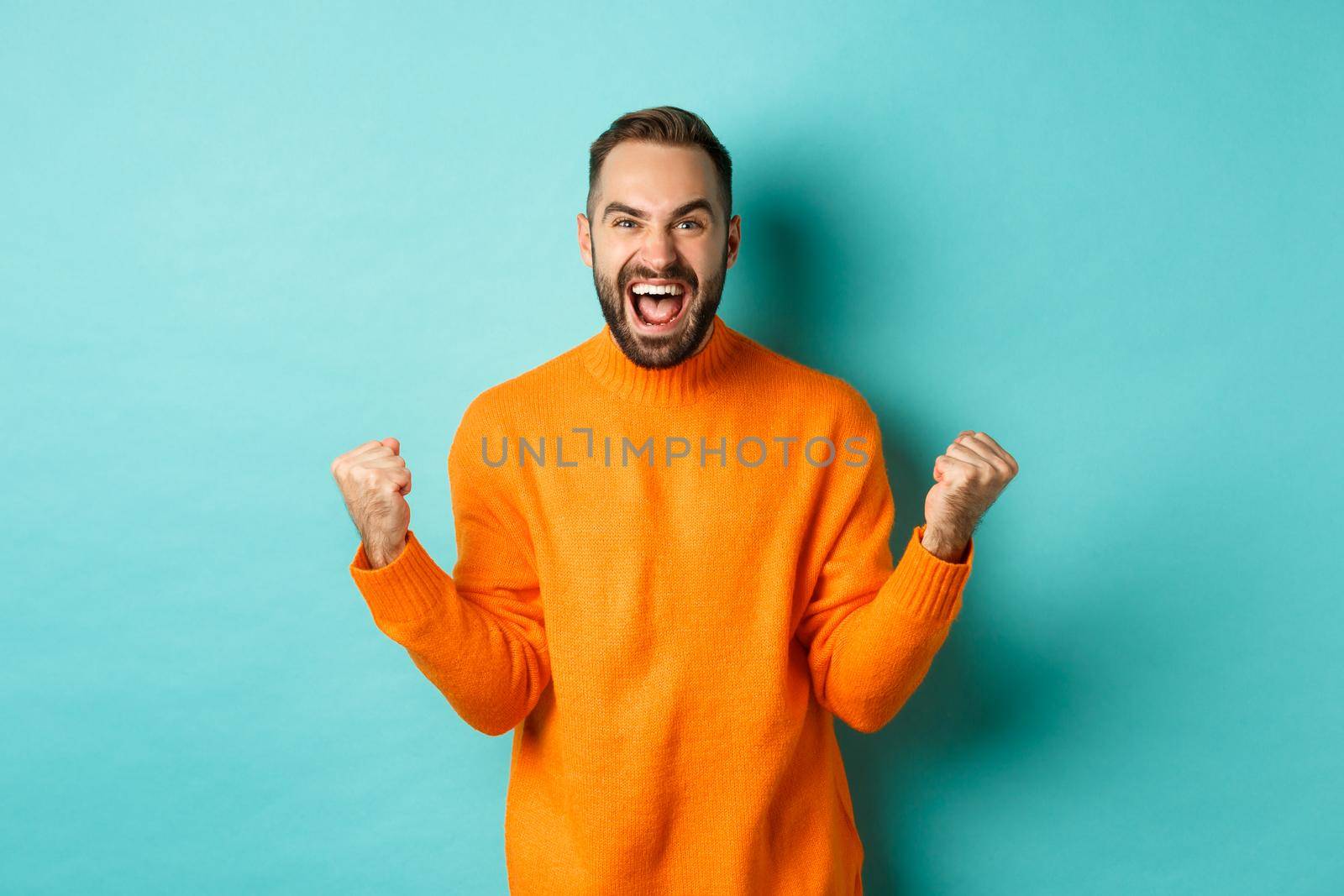 Excited man celebrating victory, rejoicing and making fist pump gesture, winning and looking satisfied, saying yes, achieve goal, standing over light blue background.