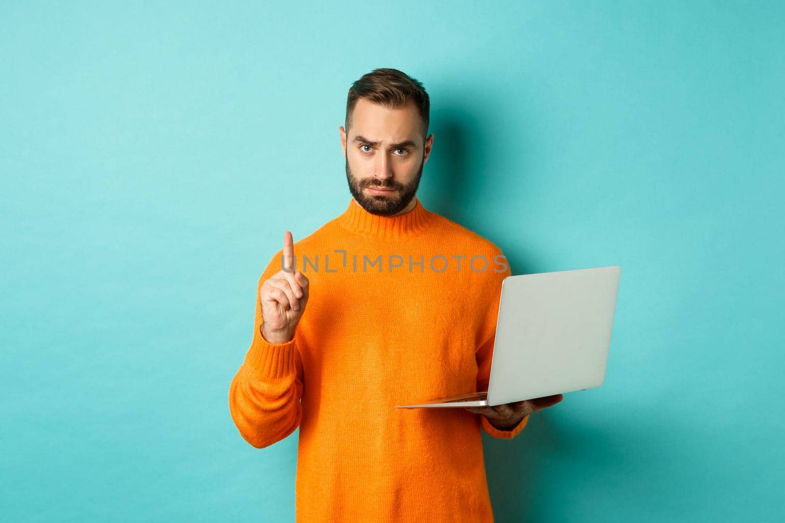 Image of adult man teaching online, showing finger while using laptop, explaining rules, standing over light blue background.