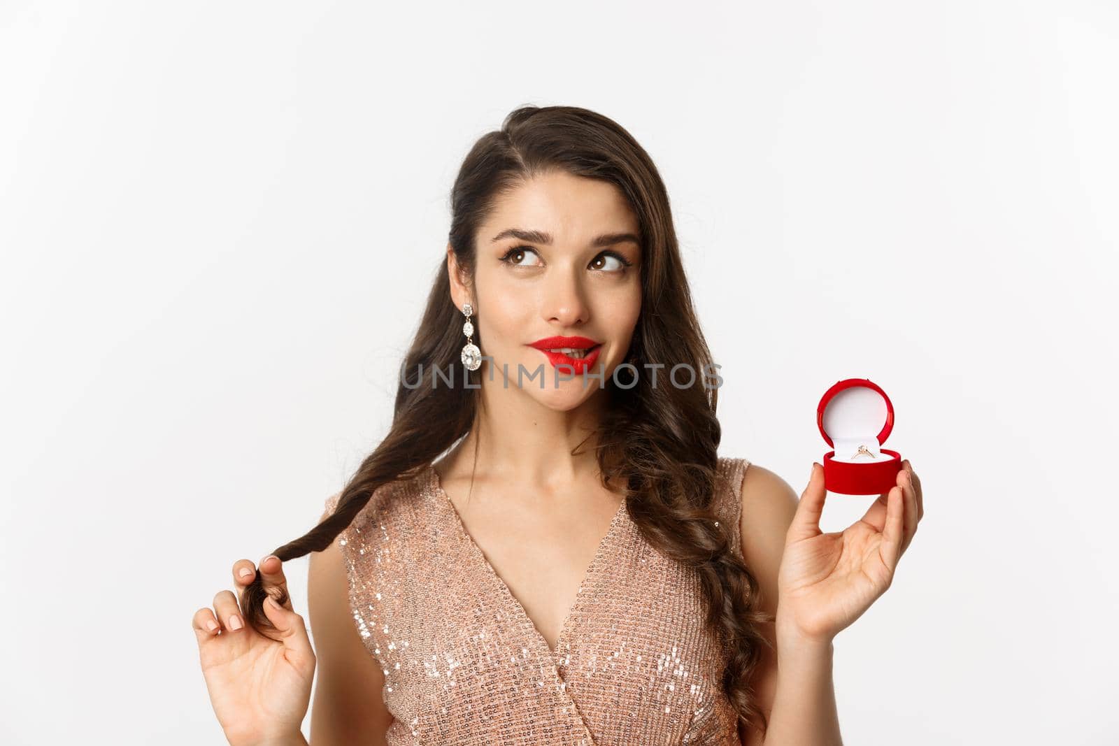 Close-up of coquettish beautiful woman thinking about marriage, holding engagement ring and looking at upper left corner, smiling silly, white background.