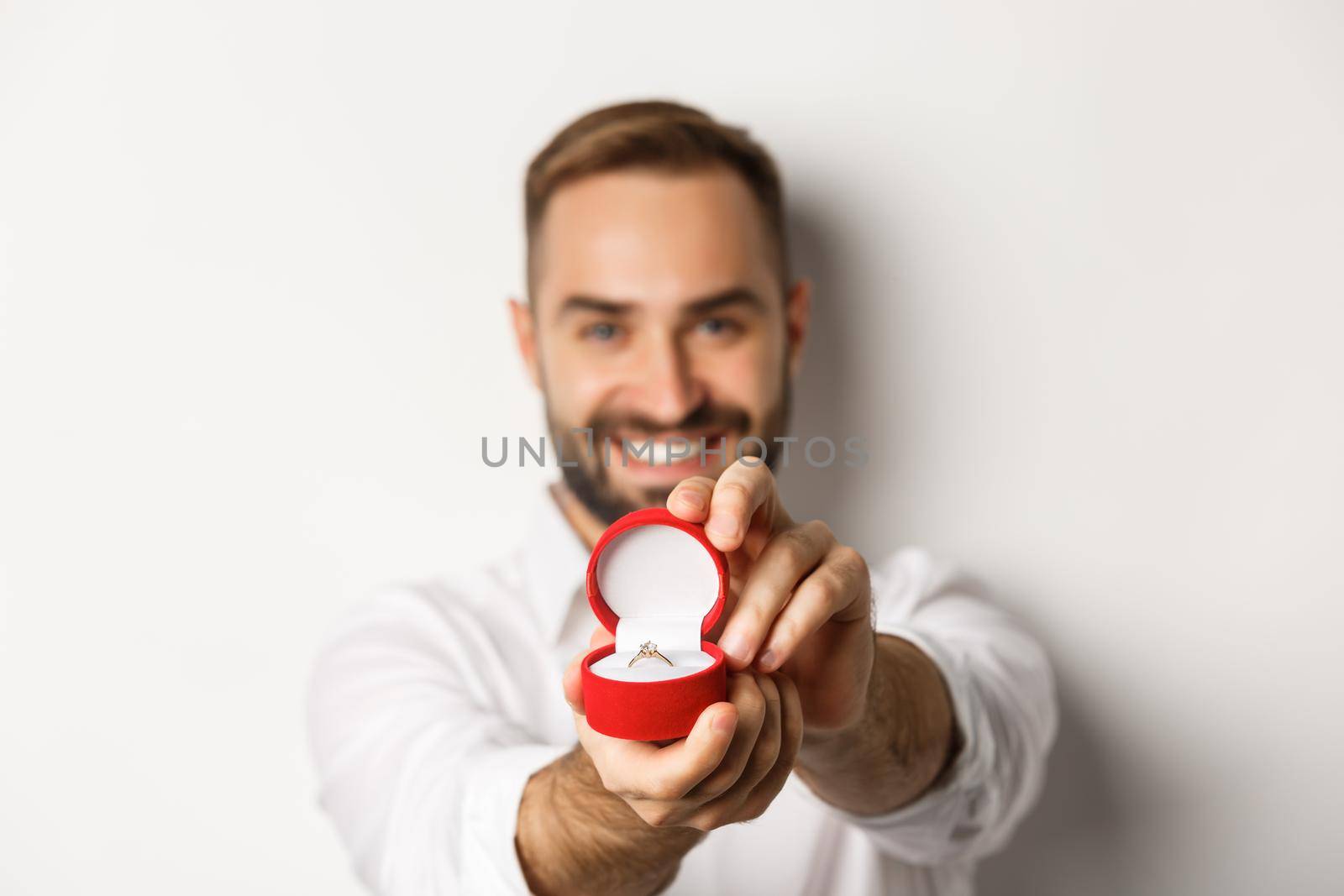Close-up of handsome man asking to marry him, focus on box with wedding ring, concept of proposal and relationship, white background.