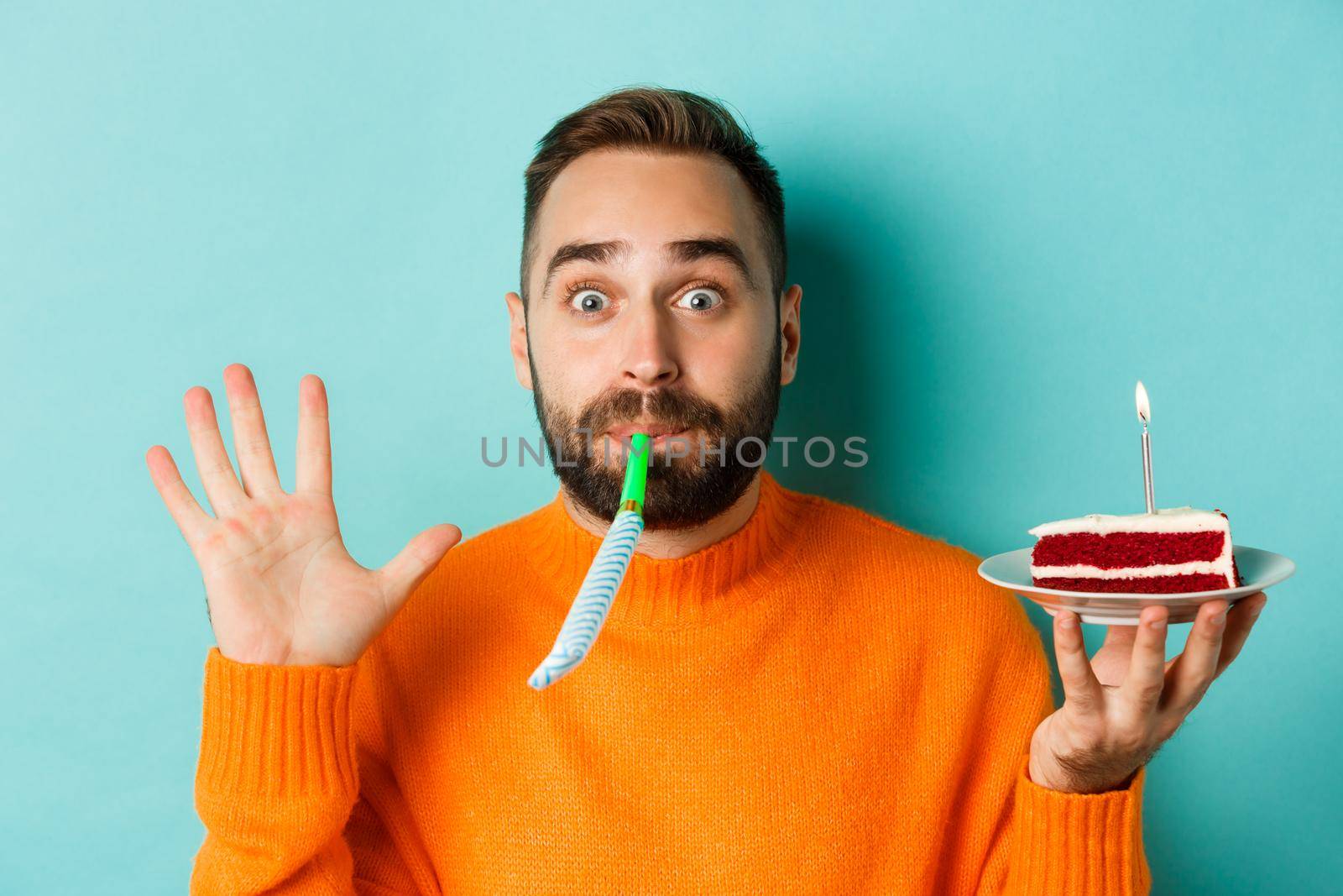 Close-up of funny adult man celebrating his birthday, holding bday cake with candle, blowing party wistle and rejoicing, standing over light blue background by Benzoix