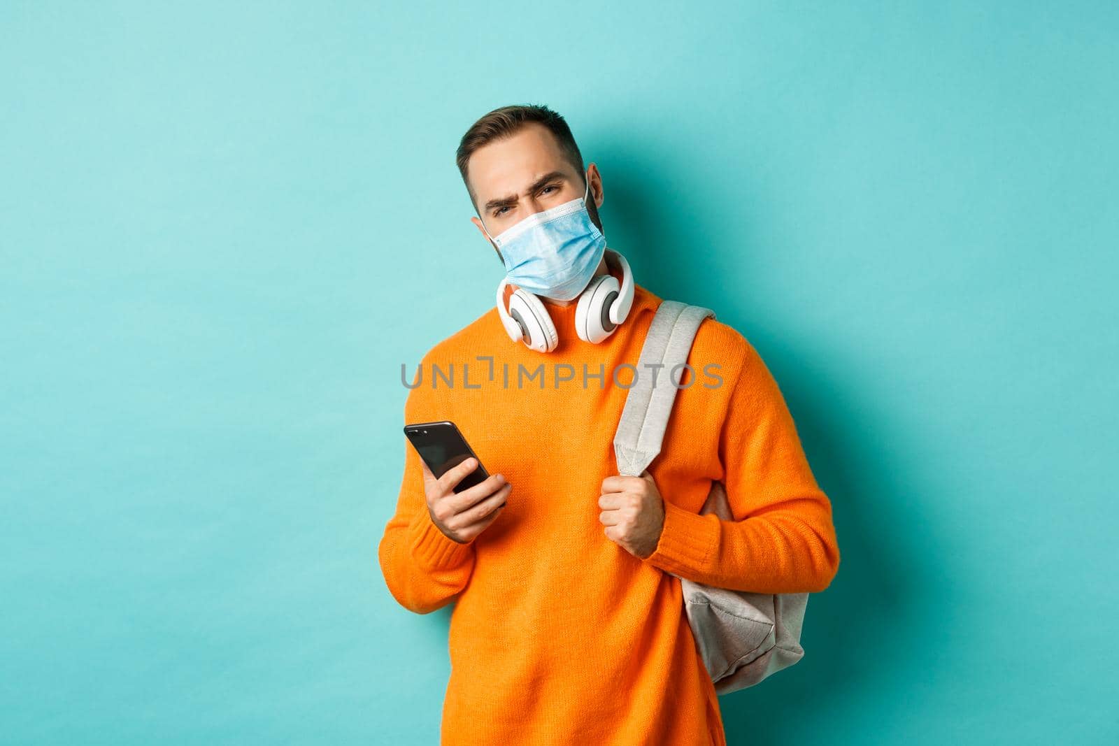 Skeptical and disappointed young man wearing face mask, holding backpack and mobile phone, frowning upset, standing over light blue background.