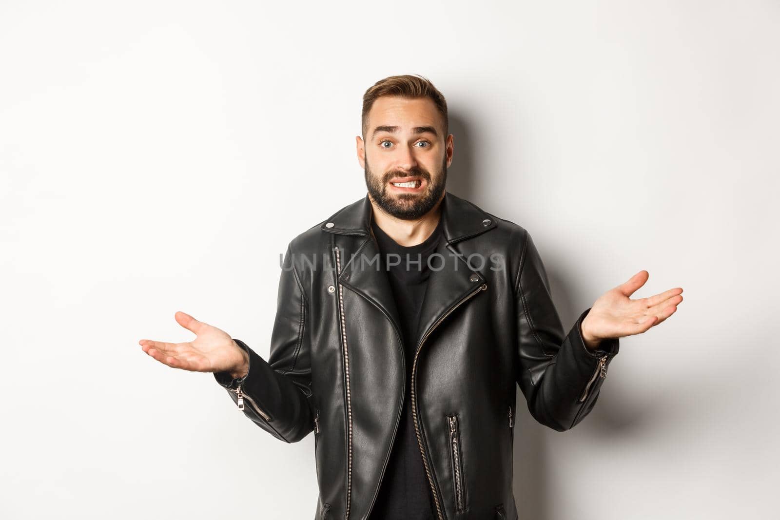Confused bearded man in black leather jacket asking for advice, looking clueless and shrugging, standing over white background.