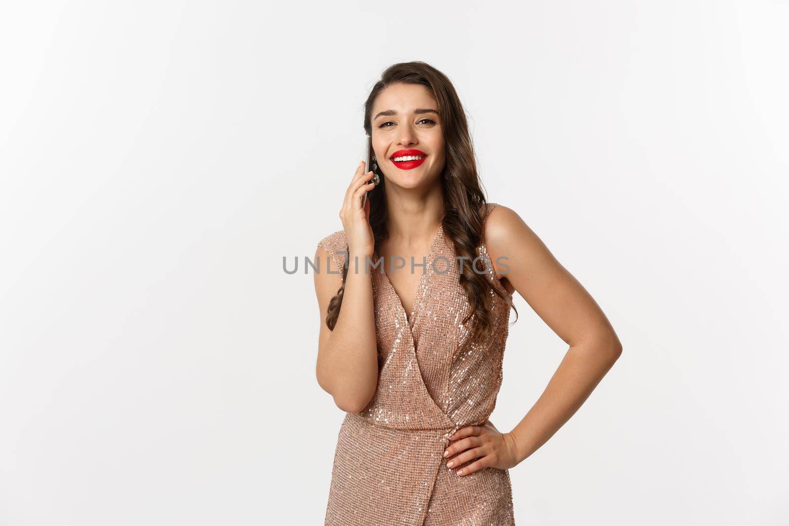 Christmas party and celebration concept. Attractive young woman with red lips, wearing luxury dress, talking on mobile phone and smiling, standing over white background by Benzoix