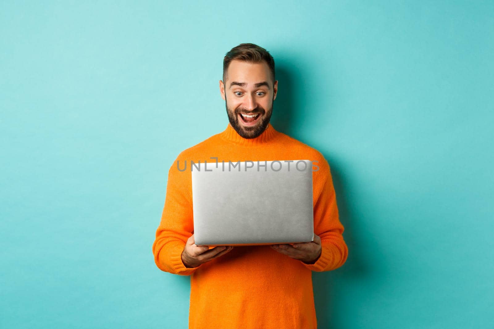 Excited man in orange sweater working on laptop, looking at computer screen amazed, standing over light blue background by Benzoix