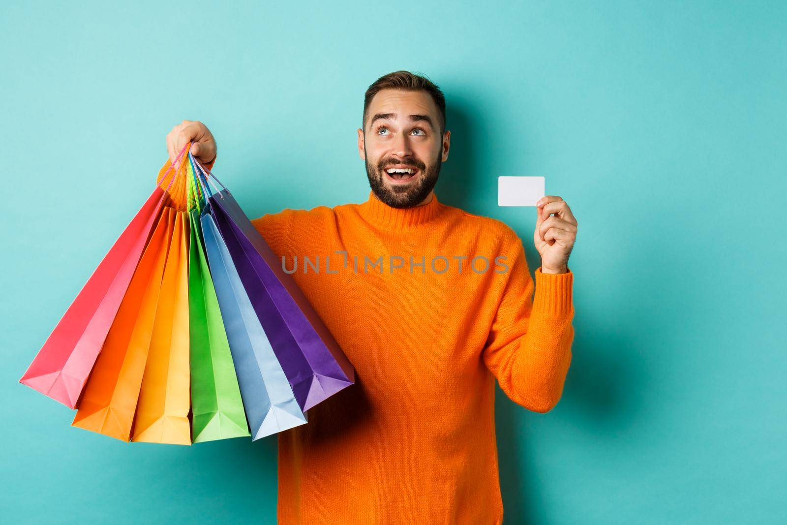Happy aduly man showing credit card and shopping bags, standing against turquoise background by Benzoix