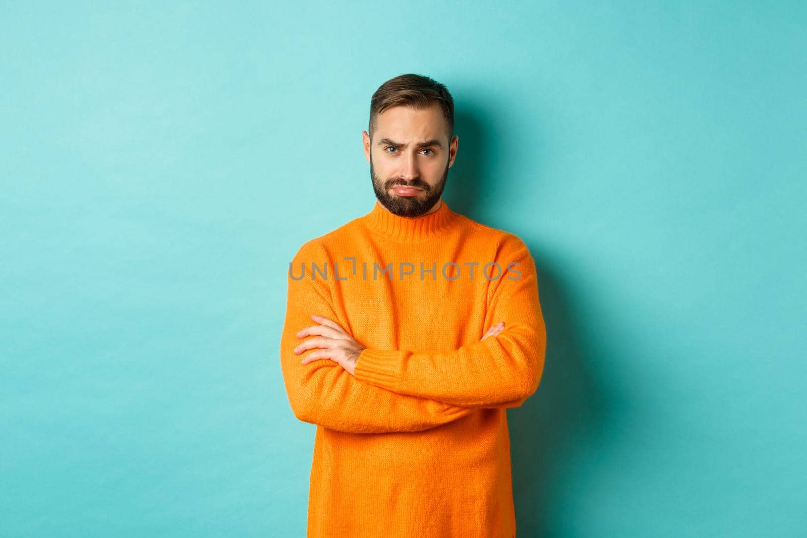 Silly sad guy with beard, sulking and looking offended, cross arms on chest and frowning upset, standing over light blue background by Benzoix