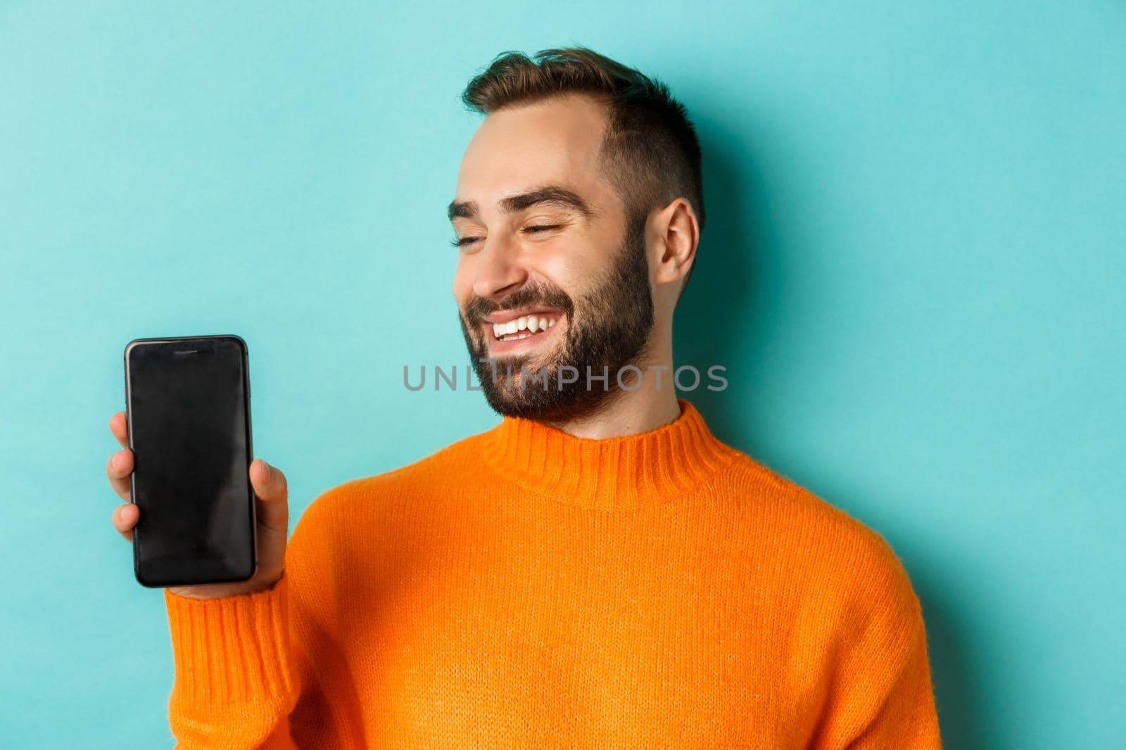 Close-up of young bearded man showing phone screen and looking satisfied, wearing orange sweater, standing against studio background by Benzoix