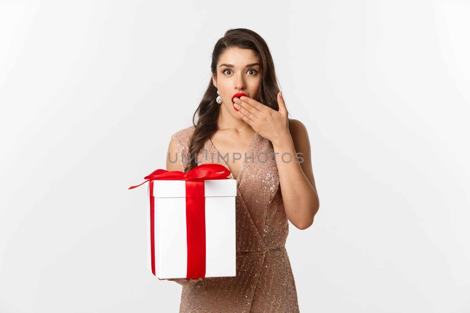 Merry Christmas. Beautiful woman looking surprised and holding gift, receiving new year presents, standing in luxury dress, standing over white background.