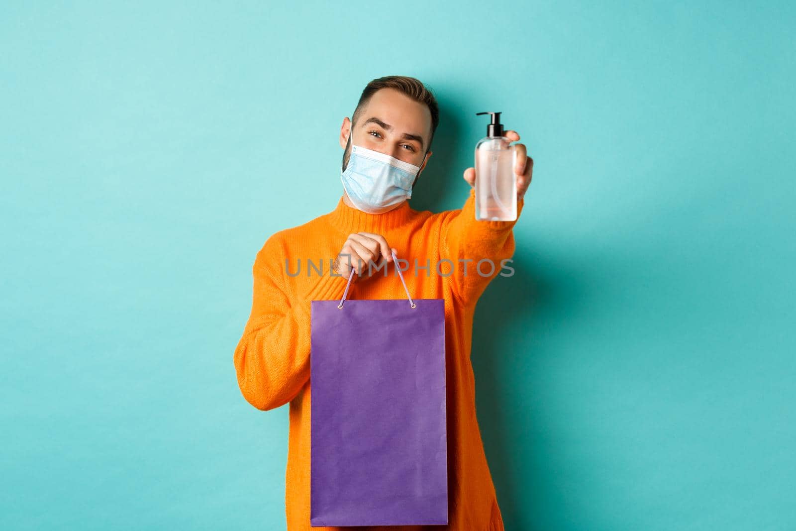 Coronavirus, pandemic and lifestyle concept. Man in face mask showing shopping bag and hand sanitizer, standing over turquoise background.