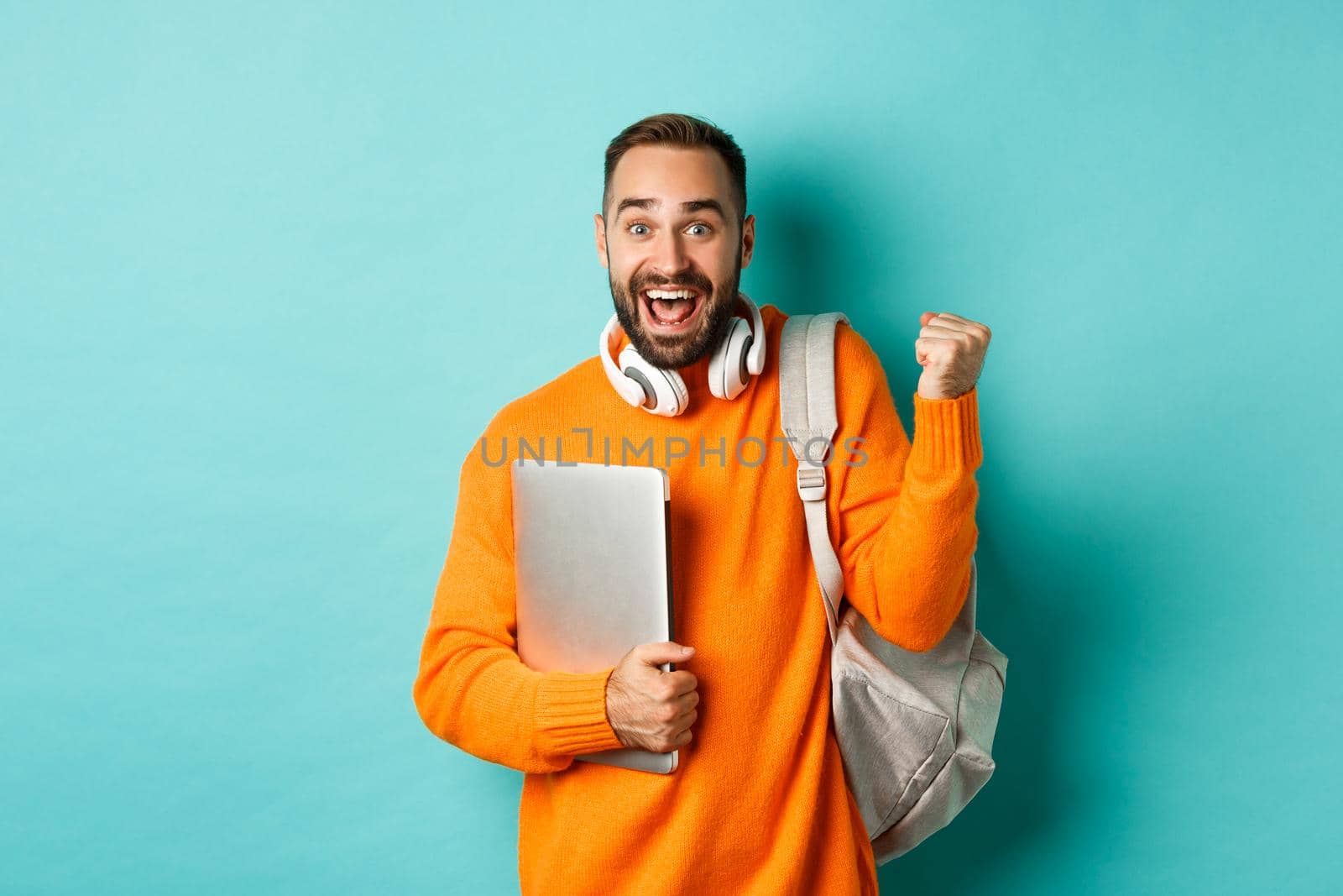 Happy man with backpack and headphones, holding laptop and smiling, cheering of win, triumphing over turquoise background.