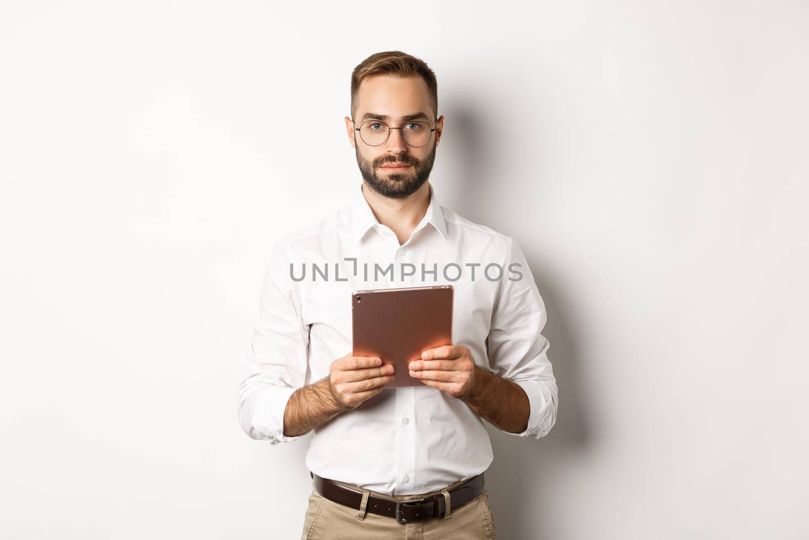 Serious employer working with digital tablet, reading in glasses, standing over white background.