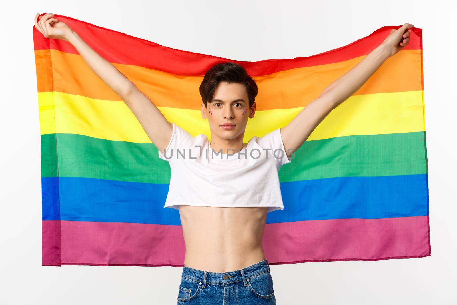 Out and proud. Happy and confident gay man with glitter on face raising lgbtq rainbow flag and smiling at camera, standing in crop top, white background by Benzoix