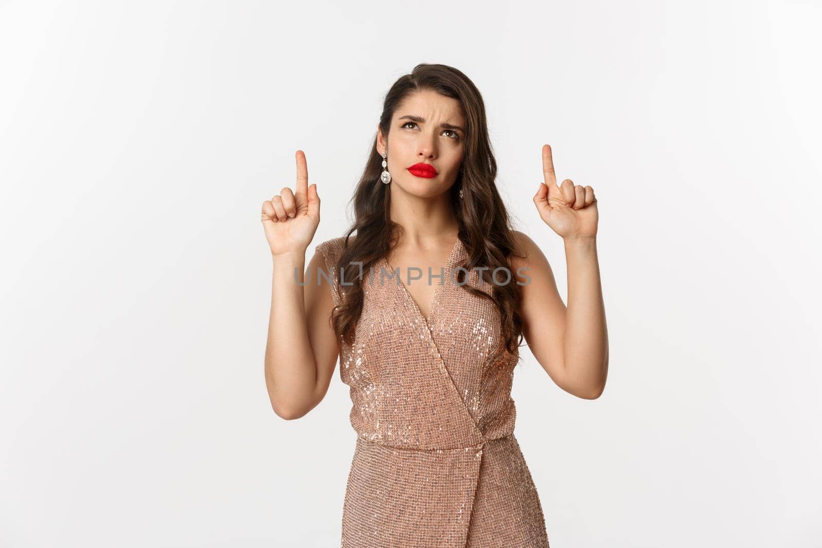 Christmas, holidays and celebration concept. Elegant woman in luxury dress looking doubtful at logo, pointing fingers up, standing over white background by Benzoix