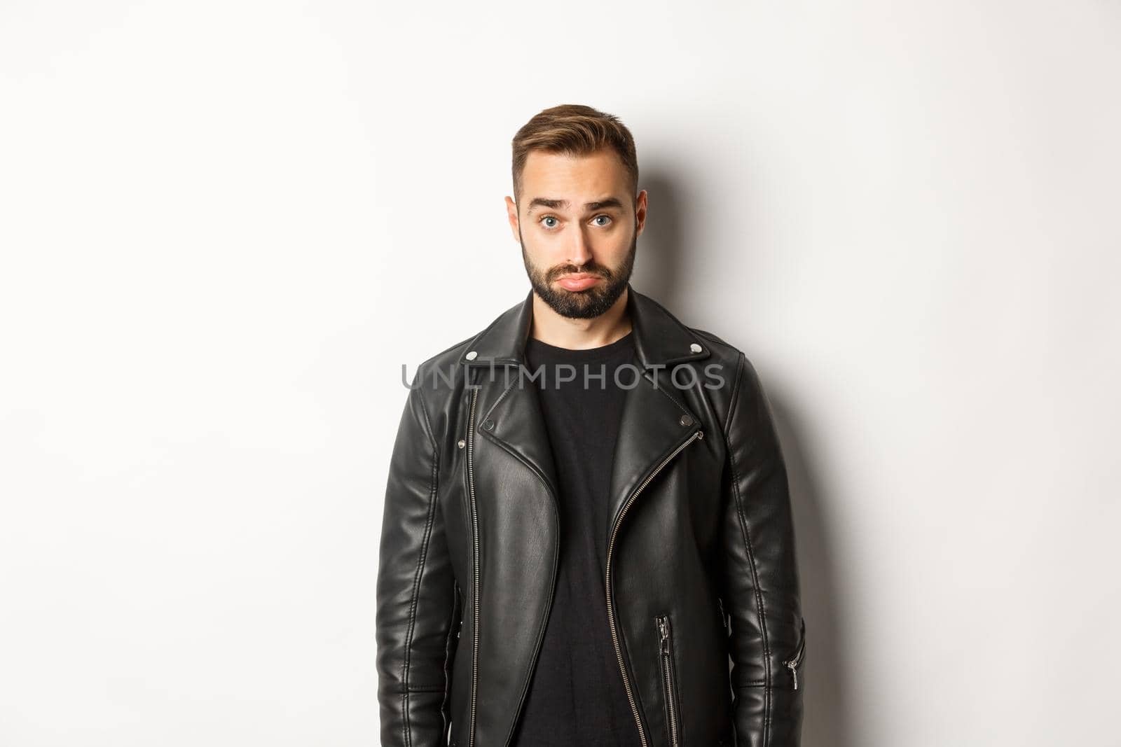 Sad and cute guy in leather black jacket pouting, looking silly at camera, standing against white background.