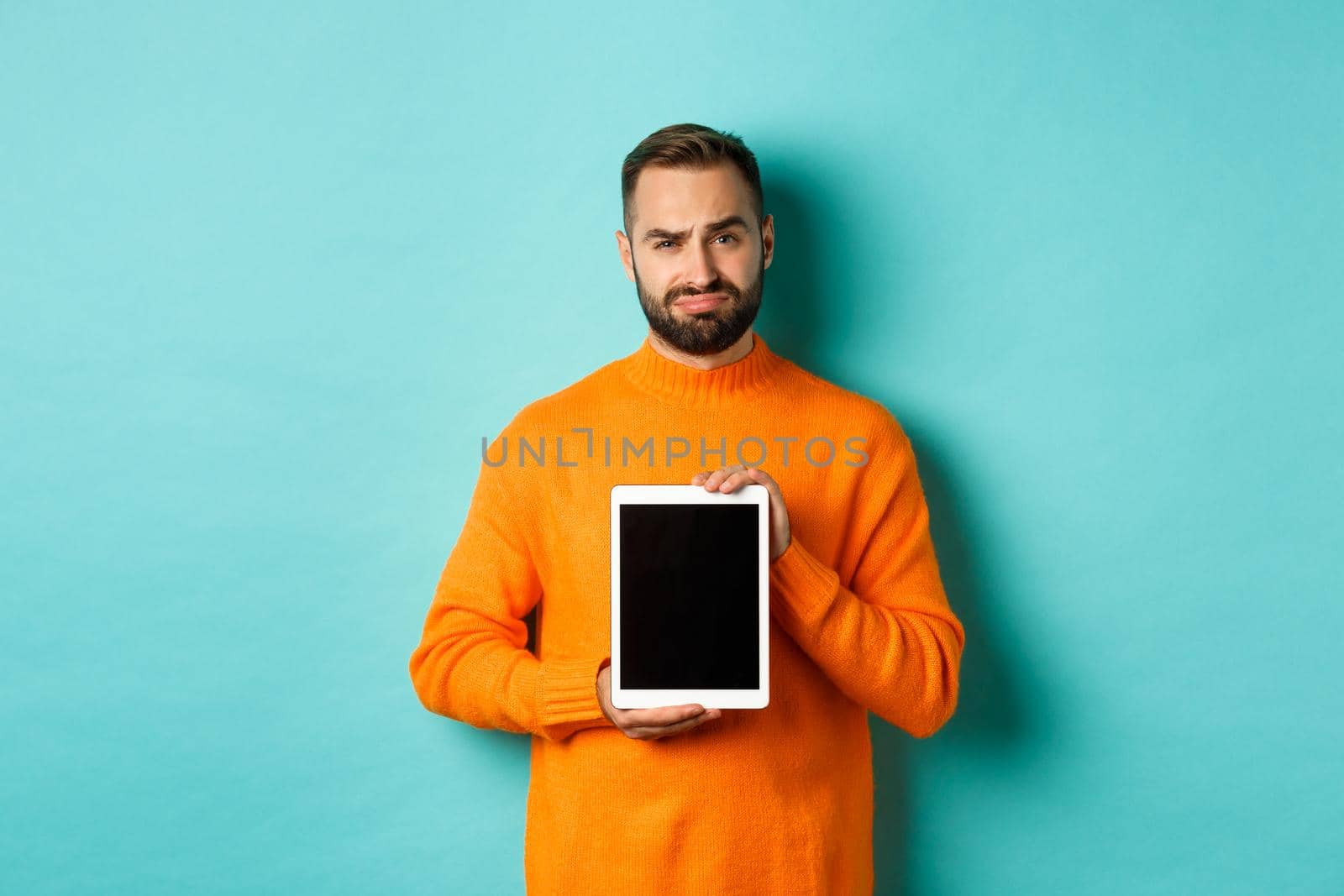 Technology. Skeptical and displeased guy showing digital tablet screen, frowning disappointed, standing over light blue background.