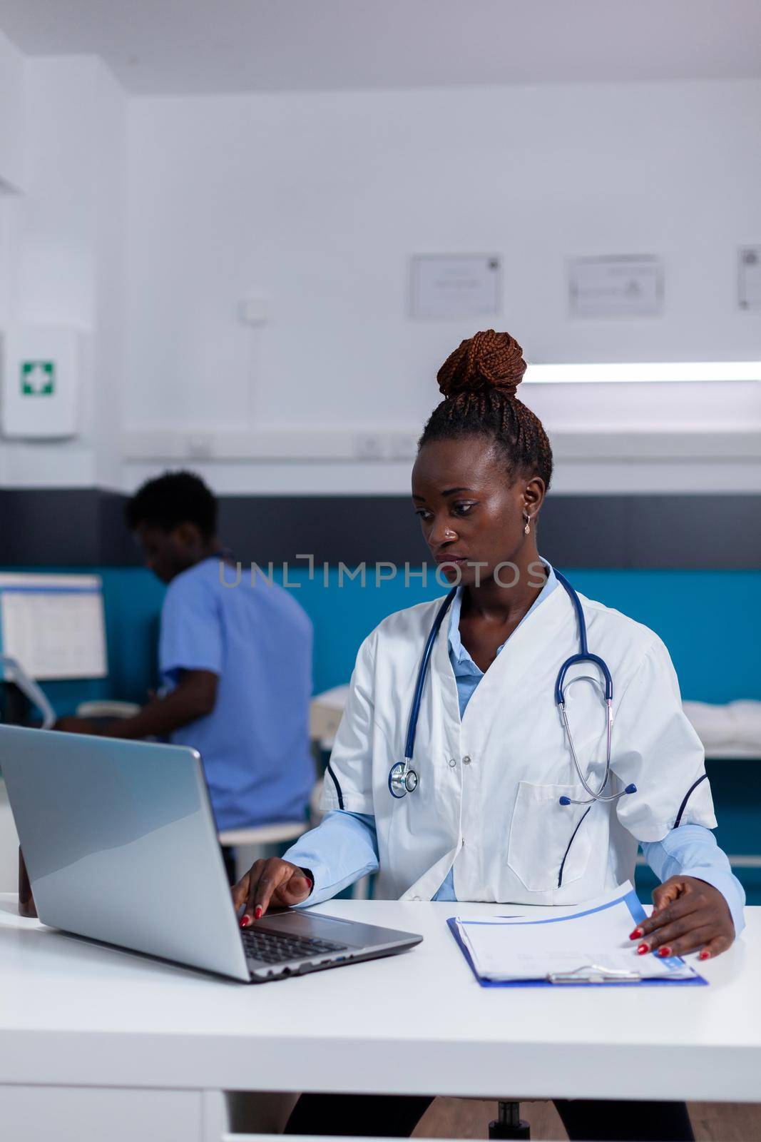 African american person with medic job using laptop at desk in medical office. Black doctor sitting with technology and healthcare equipment while nurse looking at computer in background