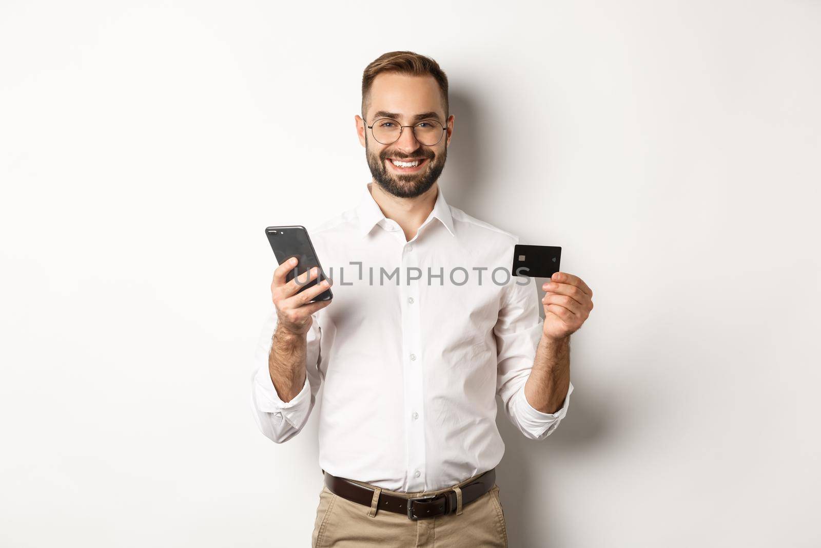 Business and online payment. Smiling male entrepreneur shopping with credit card and mobile phone, standing over white background by Benzoix