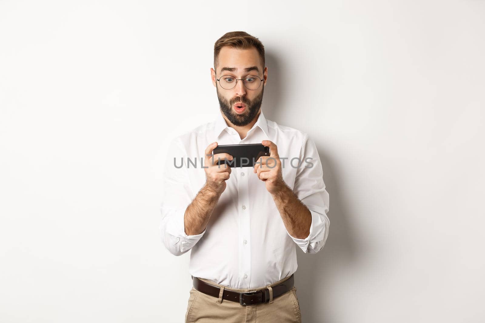 Man looking amazed at mobile phone, standing against white background.