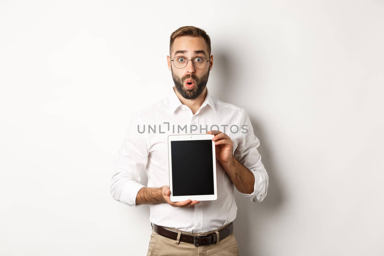 Surprised man in glasses, showing digital tablet screen, looking amazed, standing over white background.
