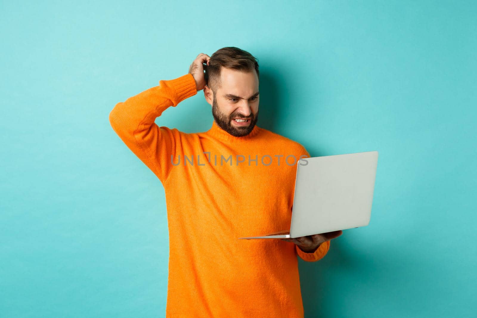 Freelance and technology concept. Confused man scratching head and looking at laptop screen puzzled, dont understand, standing over light blue background.