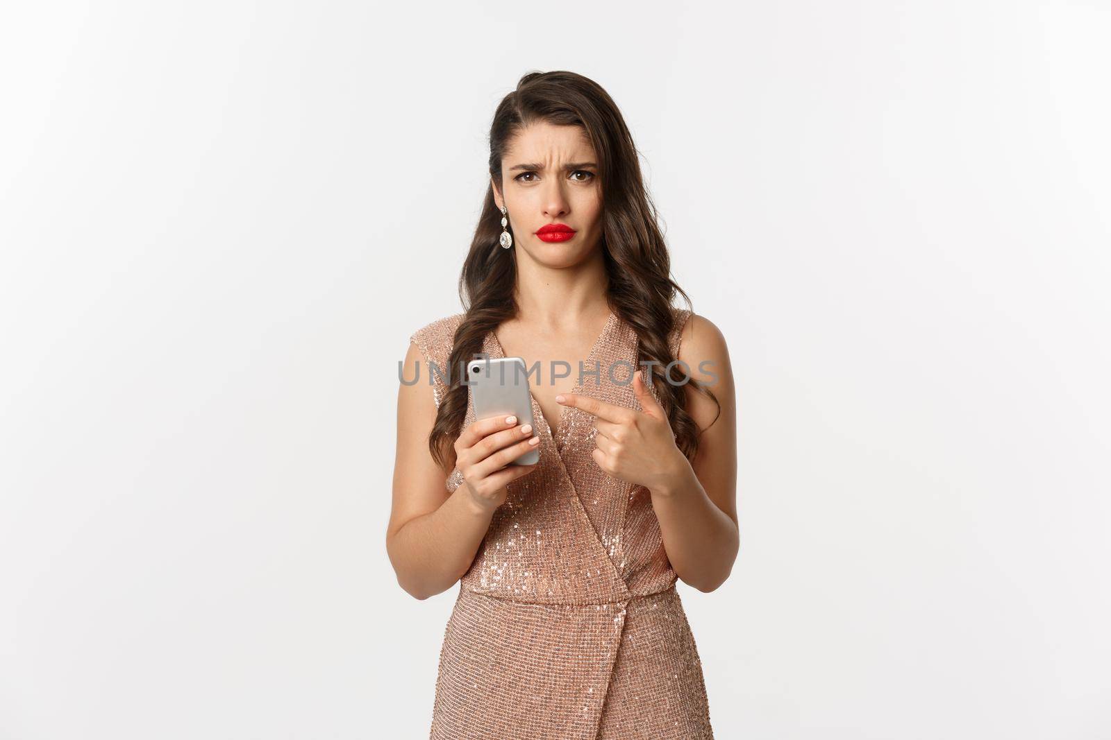 Holidays, online shopping concept. Disappointed and confused woman in elegant dress pointing at mobile phone, reacting at strange message, white background.