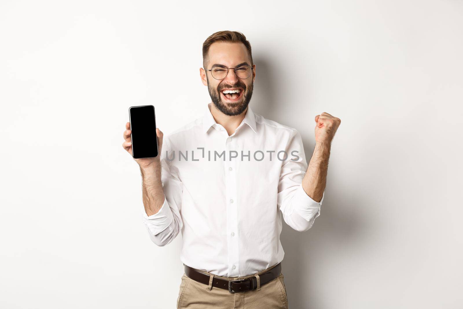 Successful business man showing mobile screen, rejoicing on winning online prize, achieve app goal, standing against white background.