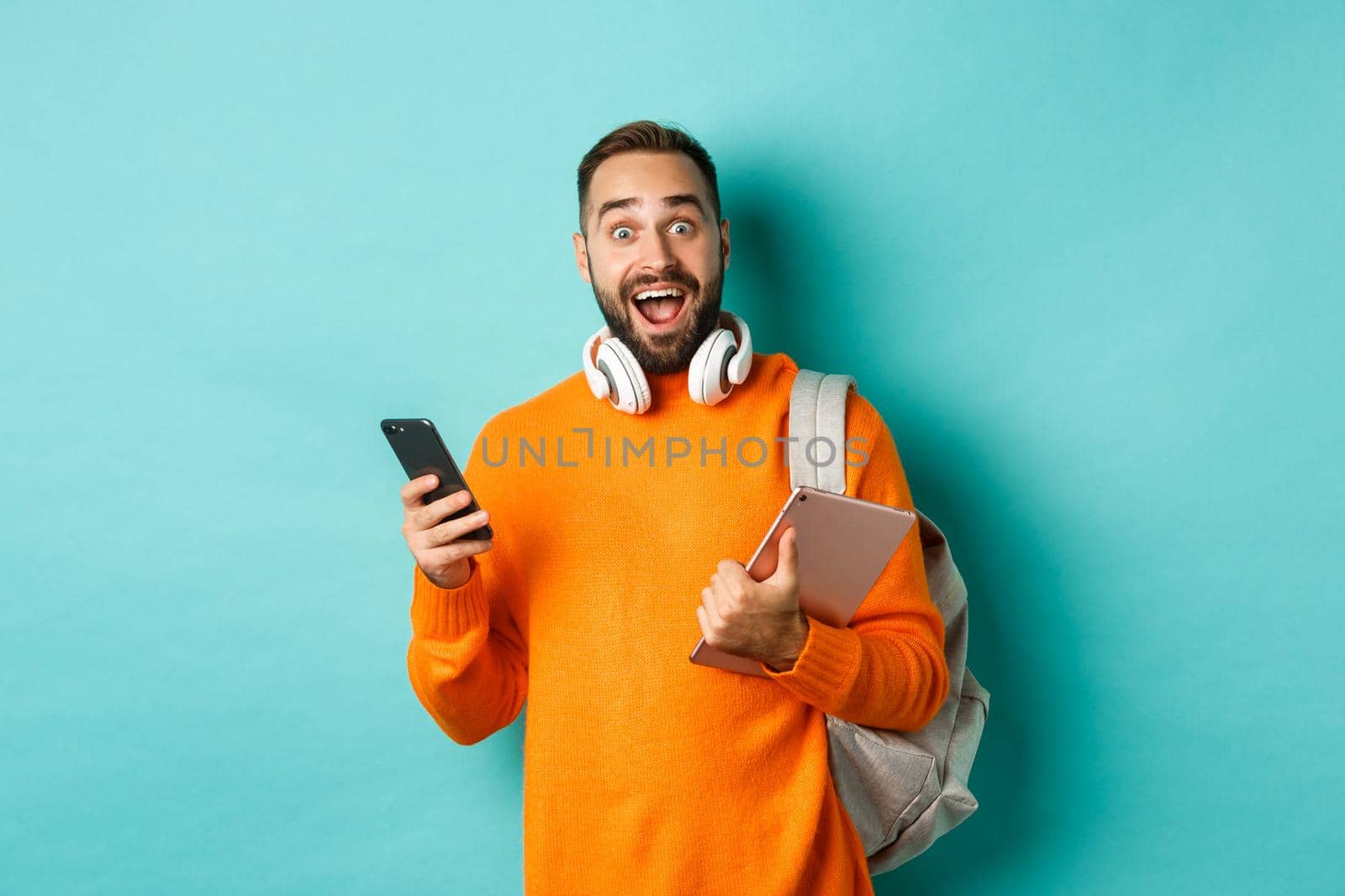 Handsome man student with headphones and backpack, holding digital tablet and smartphone, looking amazed at camera, standing against turquoise background.