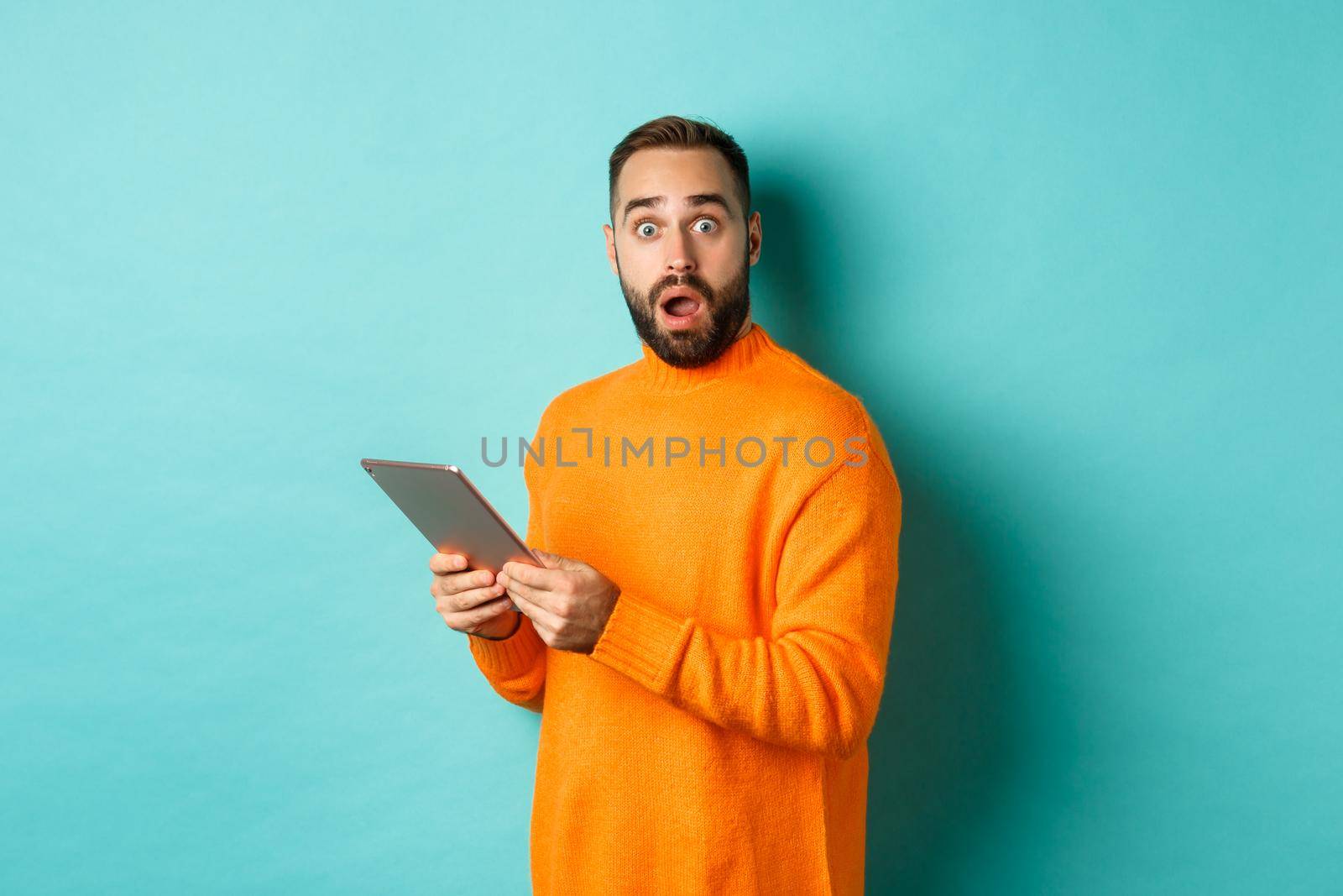 Image of male model in orange sweater using digital tablet, looking surprised, standing over light blue background.