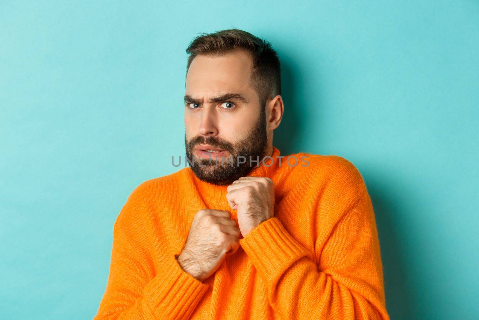 Close-up of scared bearded man looking startled and displeased, jumping from fear, standing over light blue background.