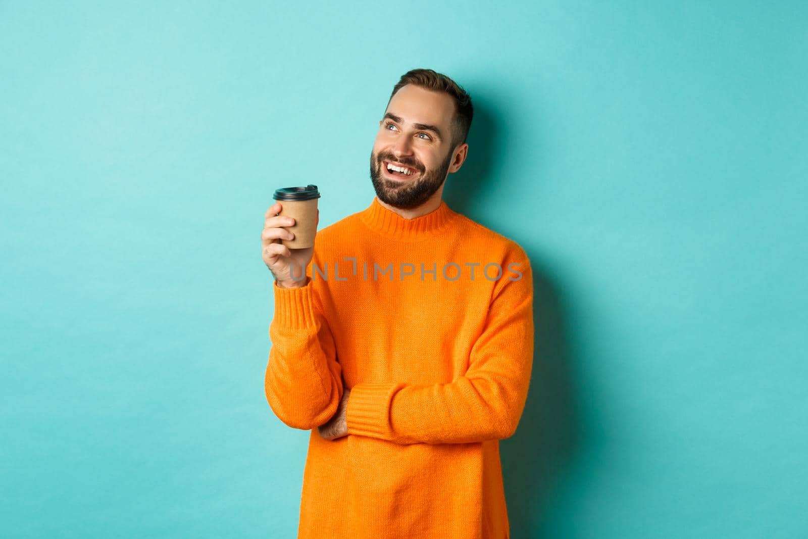 Thoughtful smiling man looking at upper left corner, having idea while drinking coffee from takeaway cup, standing in orange sweater against turquoise background.