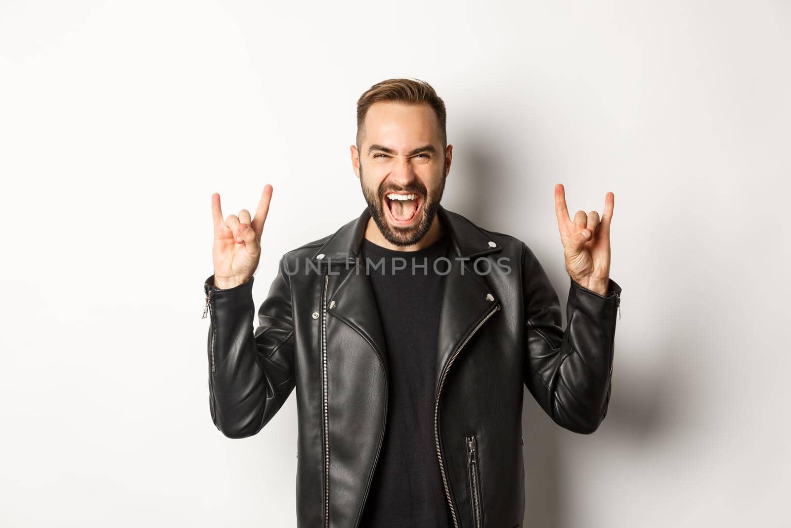 Cool adult man in black leather jacket, showing rock on gesture and tongue, enjoying music festival, standing over white background.
