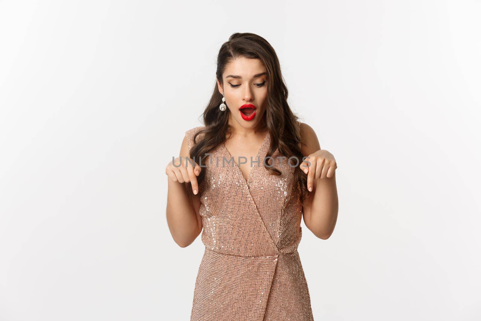 Amazed glamour girl in party dress, looking and pointing down amazed, showing christmas advertisement, standing over white background.