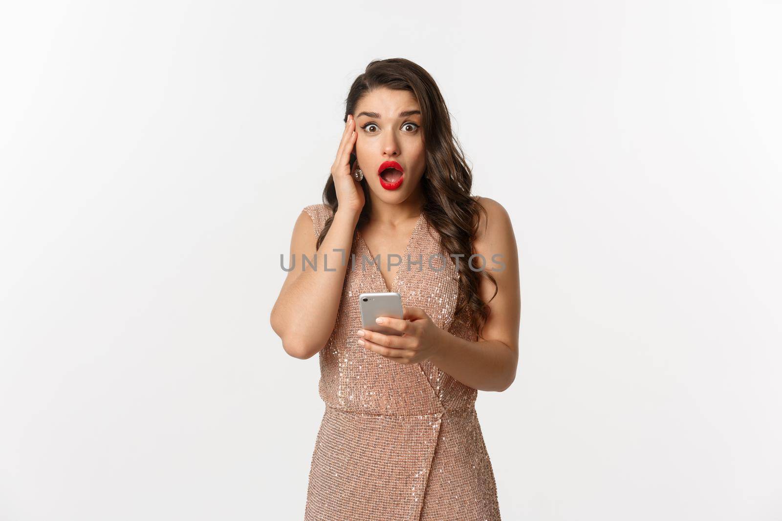 Holidays, online shopping concept. Surprised woman using mobile phone and looking amazed, wearing party dress, standing over white background.