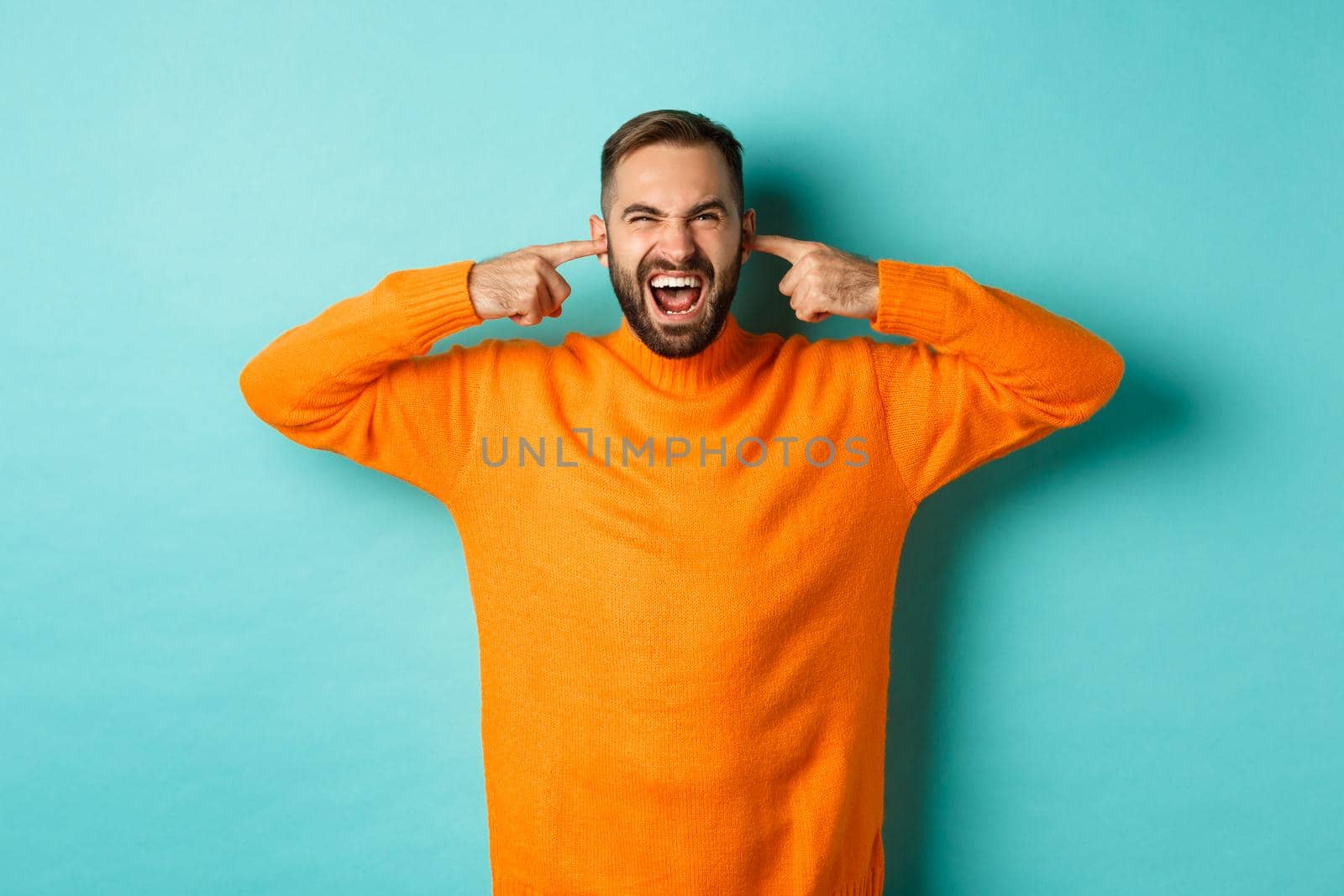 Displeased man shut ears and grimacing, loud neighbours, standing annoyed with noise, shouting angry and distressed, light blue background.