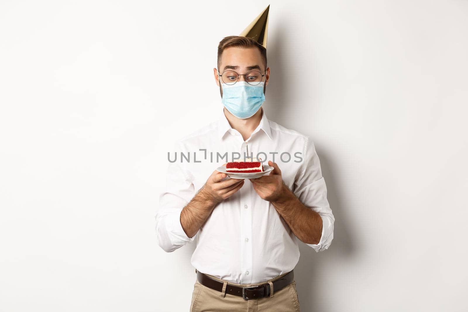 Covid-19, social distancing and celebration. Man looking excited at birthday cake, wearing medical mask from coronavirus pandemic, white background.