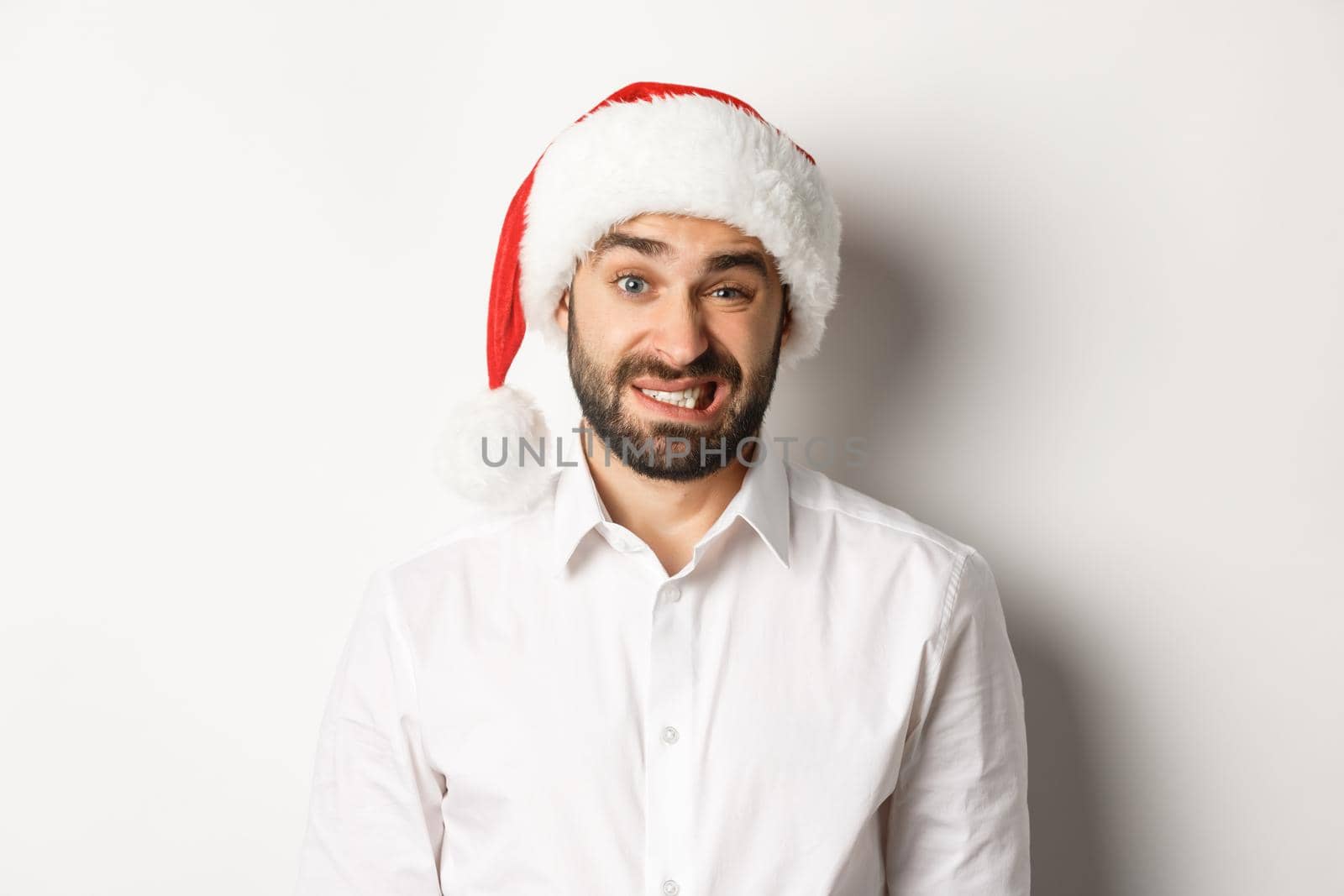 Close-up of awkward guy in santa hat apologizing, feeling uncomfortable, standing over white background. Christmas concept.