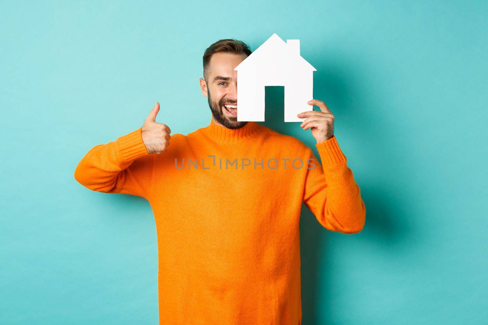 Real estate concept. Satisfied man recommending agency, showing thumb up and house maket, standing over blue background.