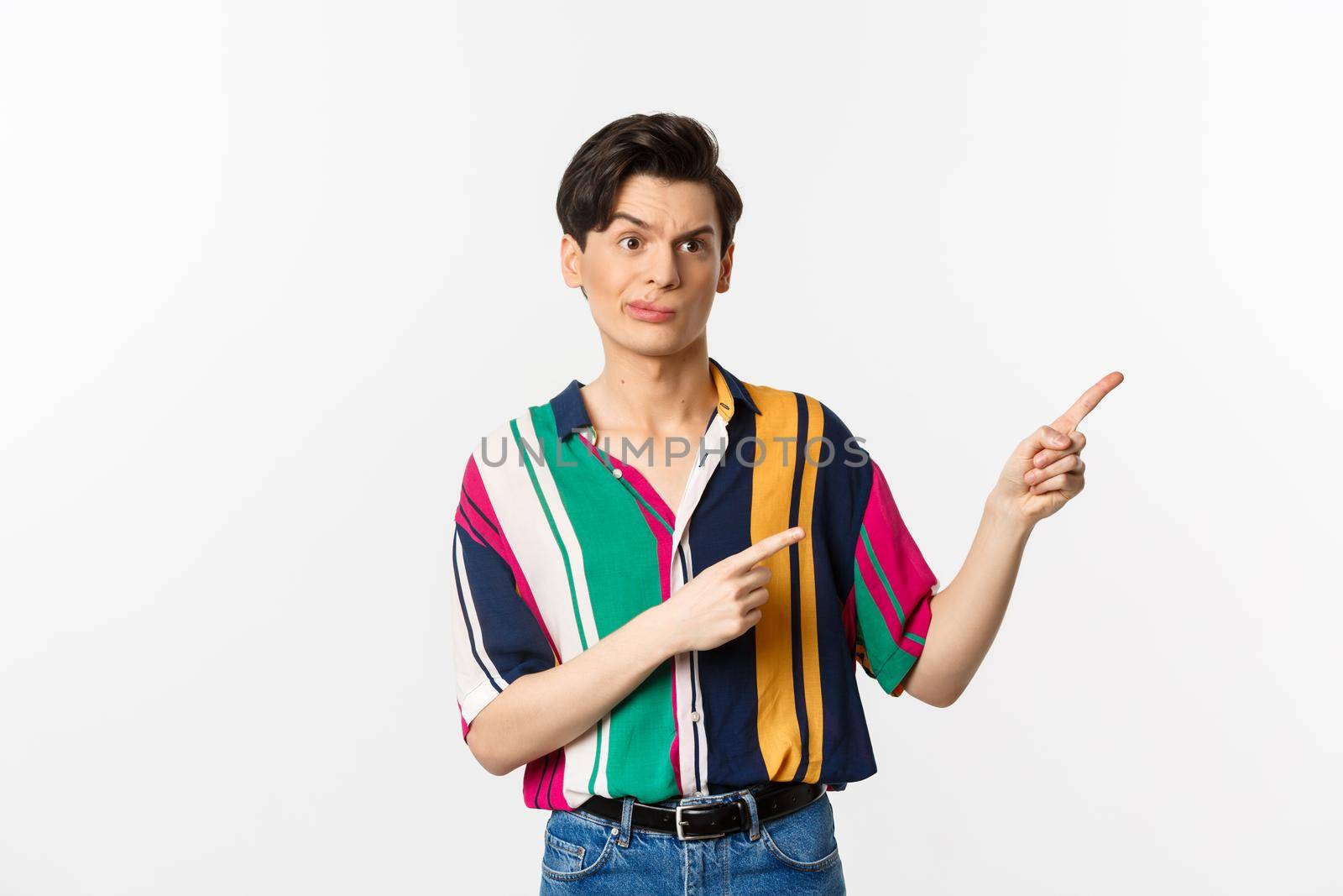 Skeptical gay man grimacing disappointed, pointing fingers right at bad product, standing over white background.